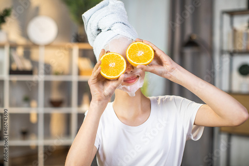 Cute female teenager hiding eyes under slices of orange while wearing vitamin mask on face. Beauty procedures for healthy and young skin. Domestic lifestyles.