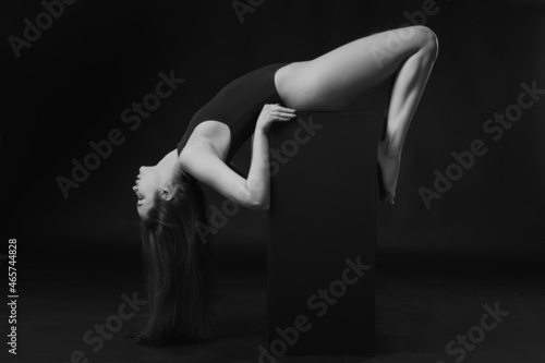 Pretty brunette woman in bodysuit posing on cube on black background. Black and white