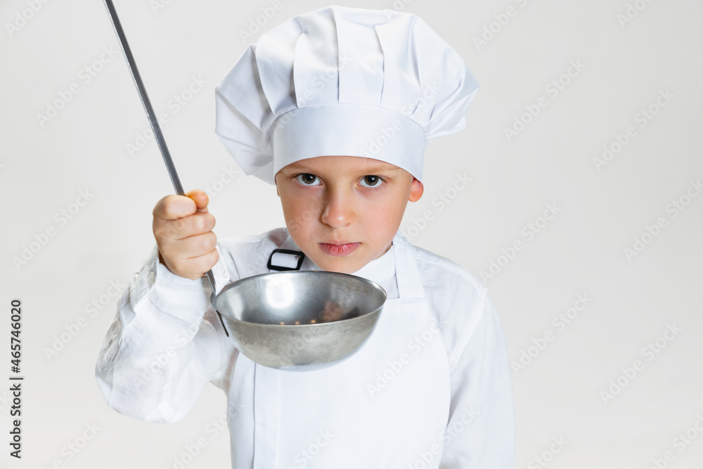 Close-up portrait of little happy boy in white cook uniform and huge chef's hat with big ladle isolated on white studio background. Close-up portrait of little happy boy in white cook uniform and huge chef's hat with big ladle isolated on white studio background.