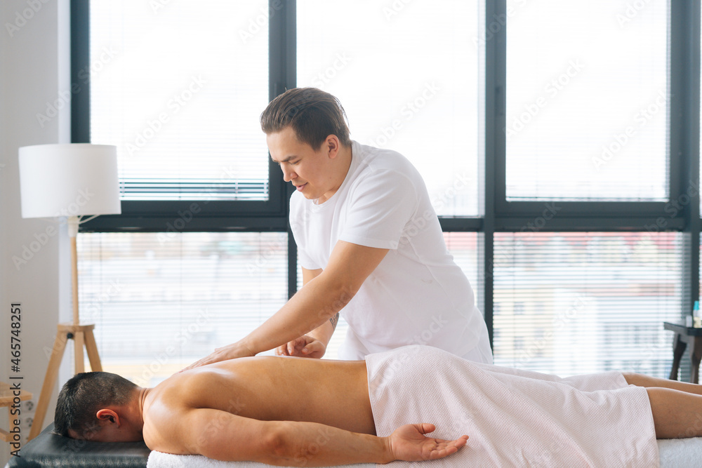 Side view of Caucasian male masseur massaging back of muscular sportsman lying on massage table on background of window. Therapist making movements with strong arms along the spine toward head. Side view of Caucasian male masseur massaging back of muscular sportsman lying on massage table on background of window. Therapist making movements with strong arms along the spine toward head.