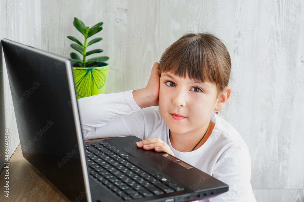 Indoor shot of cute dark haired female child sitting at table near notebook looking at camera keeping one hand on face Indoor shot of cute dark haired female child sitting at table near notebook looking at camera keeping one hand on face