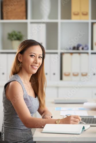 Portrait of smiling young Asian businesswoman with red hair sitting at desk in office