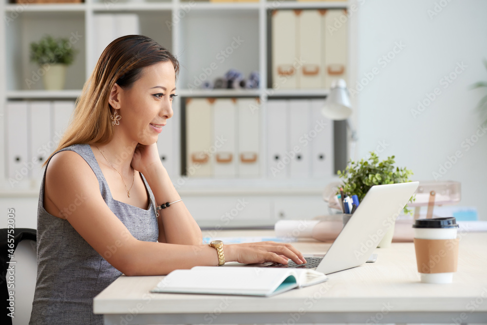 Content young Vietnamese businesswoman sitting at table in office and analyzing information on laptop