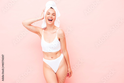 Young beautiful smiling woman in white lingerie. Sexy carefree model in underwear and towel on head posing near pink wall in studio. Positive and happy female enjoying morning