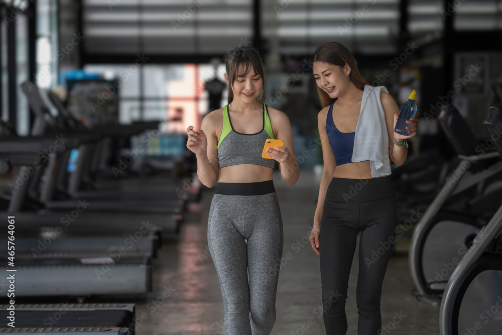 Obraz premium A smiling fit lady is showing her female friend something on phone. Both girls are resting after training together in the sports center.