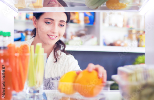 Portrait of female standing near open fridge full of healthy food, vegetables and fruits