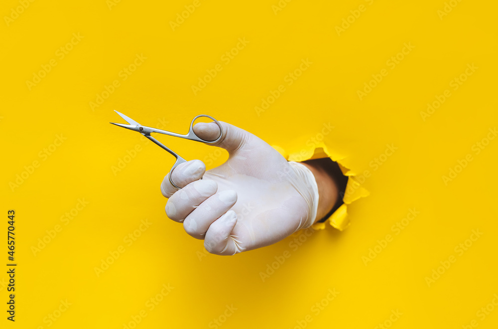 A man's hand in a white medical glove holds small scissors for a pedicure and manicure.Yellow paper background with a torn hole in yellow paper.The concept of a master nails of people and animals A man's hand in a white medical glove holds small scissors for a pedicure and manicure.Yellow paper background with a torn hole in yellow paper.The concept of a master nails of people and animals