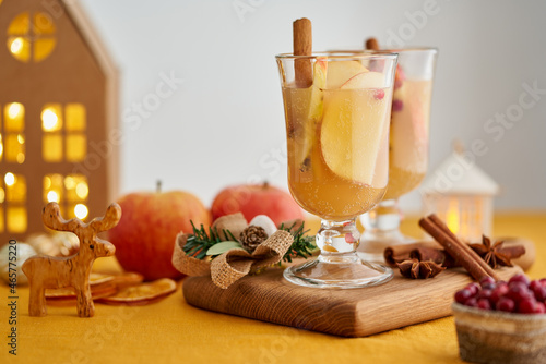 Two glasses of apple white mulled wine on wooden coaster. Christmas beverage with cinnamon, fruits and cranberry. Selective focus, blurred background, bokeh, horizontal, copy space