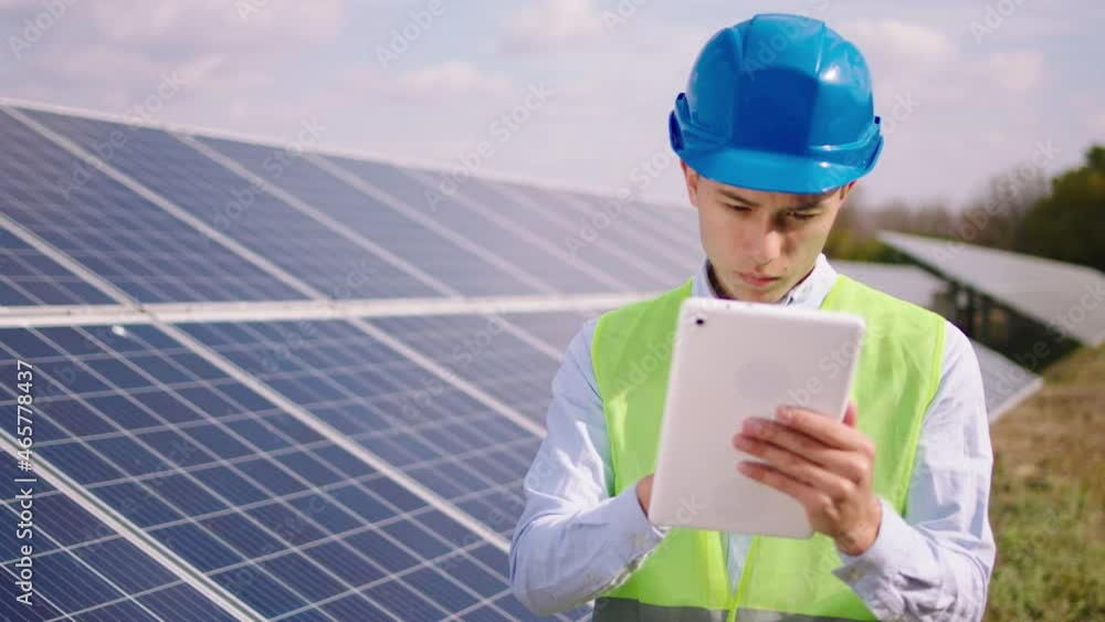 In front of the camera young ecological engineer with safety helmet at solar panels power station using digital tablet to controlling the photovoltaic batteries from wireless In front of the camera young ecological engineer with safety helmet at solar panels power station using digital tablet to controlling the photovoltaic batteries from wireless
