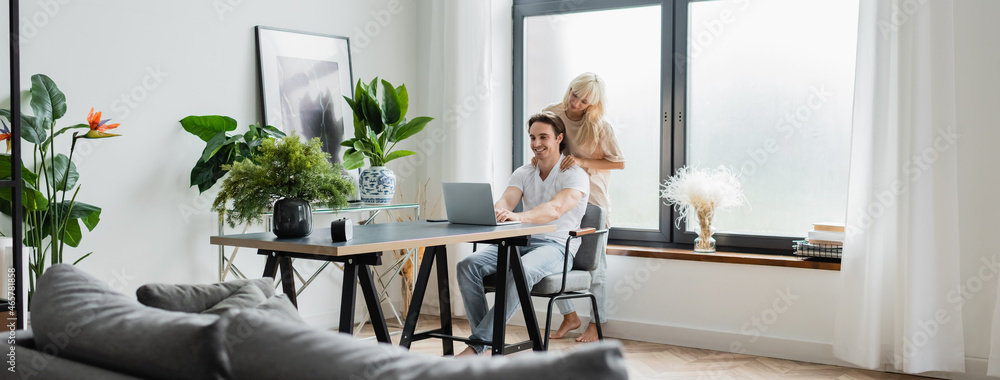 blonde woman massaging shoulders of boyfriend working from home with laptop, banner - obrazy, fototapety, plakaty 