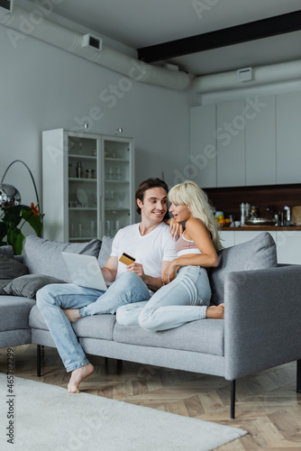 cheerful man holding credit card near amazed girlfriend while doing online shopping