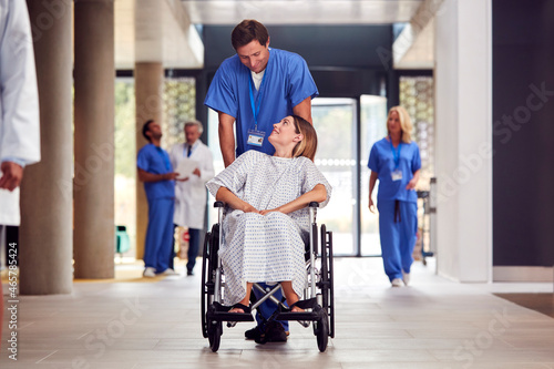 Male Nurse Wearing Scrubs Pushing Female Patient In Wheelchair Through Hospital Building