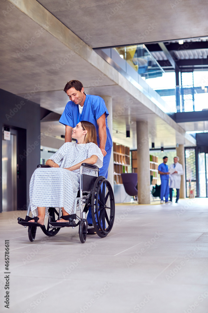 Male Nurse Wearing Scrubs Pushing Female Patient In Wheelchair Through Hospital Building Male Nurse Wearing Scrubs Pushing Female Patient In Wheelchair Through Hospital Building