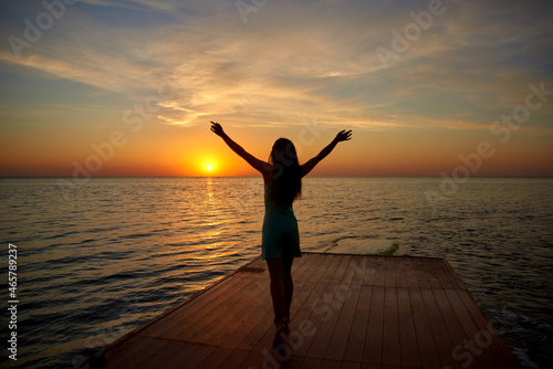 silhouette girl standing on pier raising her hands to sun. sunrise or dawn on sea or ocean side. shallow depth of field