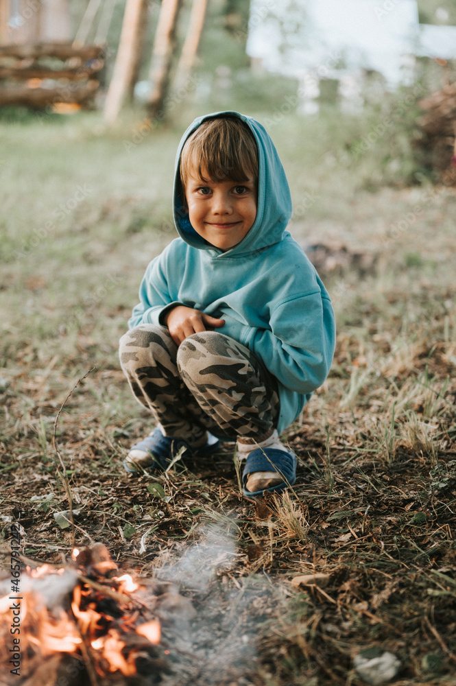 cute little happy five year old kid boy in a blue hoodie kindles a campfire in the village or forest on nature outdoor on a summer evening. authentic countryside and children's rustic life cute little happy five year old kid boy in a blue hoodie kindles a campfire in the village or forest on nature outdoor on a summer evening. authentic countryside and children's rustic life