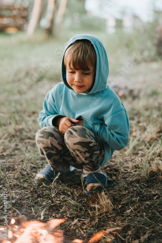 cute little happy five year old kid boy in a blue hoodie kindles a campfire in the village or forest on nature outdoor on a summer evening. authentic countryside and children's rustic life