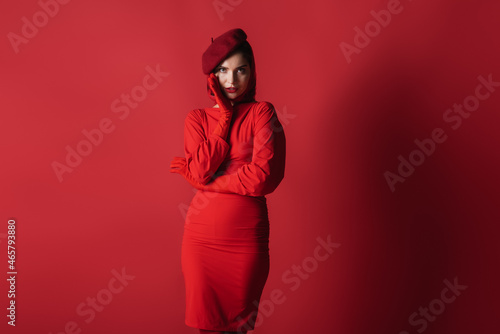 young elegant woman in beret and dress posing while looking at camera on red.