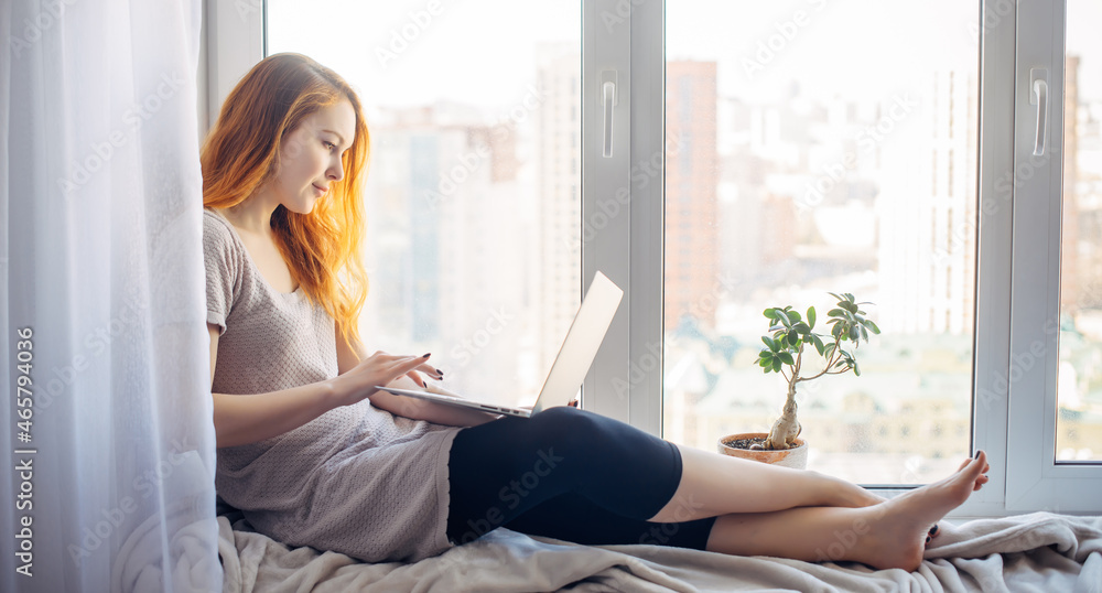 Young attractive brunette sitting on the windowsill, with laptop on her lap, typing on keyboard. Beautiful long-haired woman works at home, in city apartment. Young attractive brunette sitting on the windowsill, with laptop on her lap, typing on keyboard. Beautiful long-haired woman works at home, in city apartment.