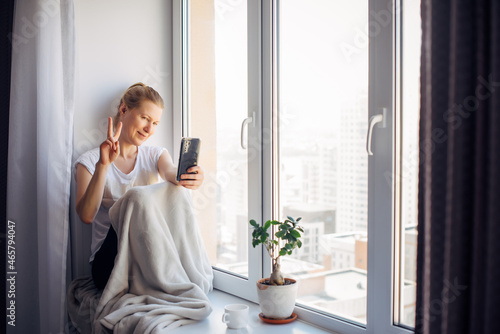Young adult woman with glasses takes a selfie sitting on the windowsill at home, gesturing with her hand. Female smiles while chatting, looking at smartphone.