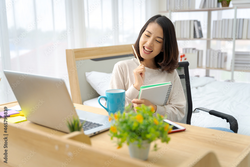 Asian woman sitting at home working in the bedroom take notes of the meeting Record work with a notebook. Cheerful, fun at work. Asian woman sitting at home working in the bedroom take notes of the meeting Record work with a notebook. Cheerful, fun at work.