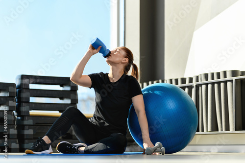 Woman with the Exercise Ball, drinks water from a bottle in the gym. Middle-aged Fitness Woman. Sport and healthy lifestyle concepts