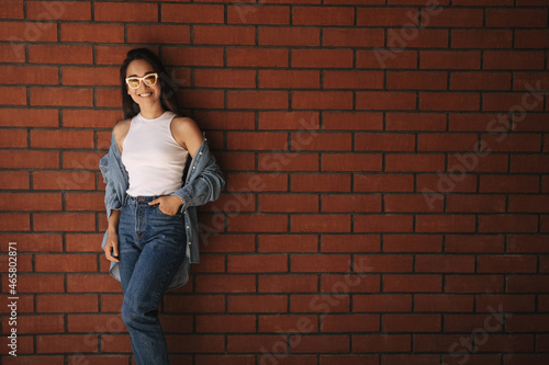 Cute slender young asian woman stands beautifully against the wall with full-length hand in her pocket. She is wearing white T-shirt, blue shirt and jeans. Mood, lifestyle, concept