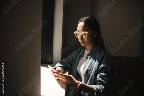 Close-up of serious Asian woman working on phone in her spare time in dark room by window. Girl with gathered hair in ponytail is wearing white top and denim shirt on top. photo