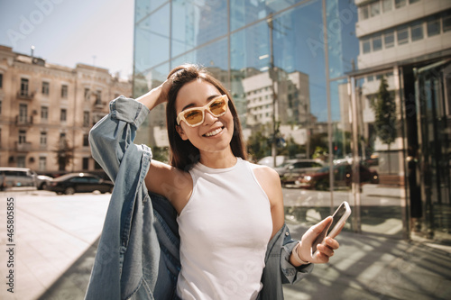 Pretty young asian girl posing in sunglasses and phone in her hands. Beauty with snow-white smile is dressed in light, light-colored casual clothes. Relaxed freelance lifestyle, concept.