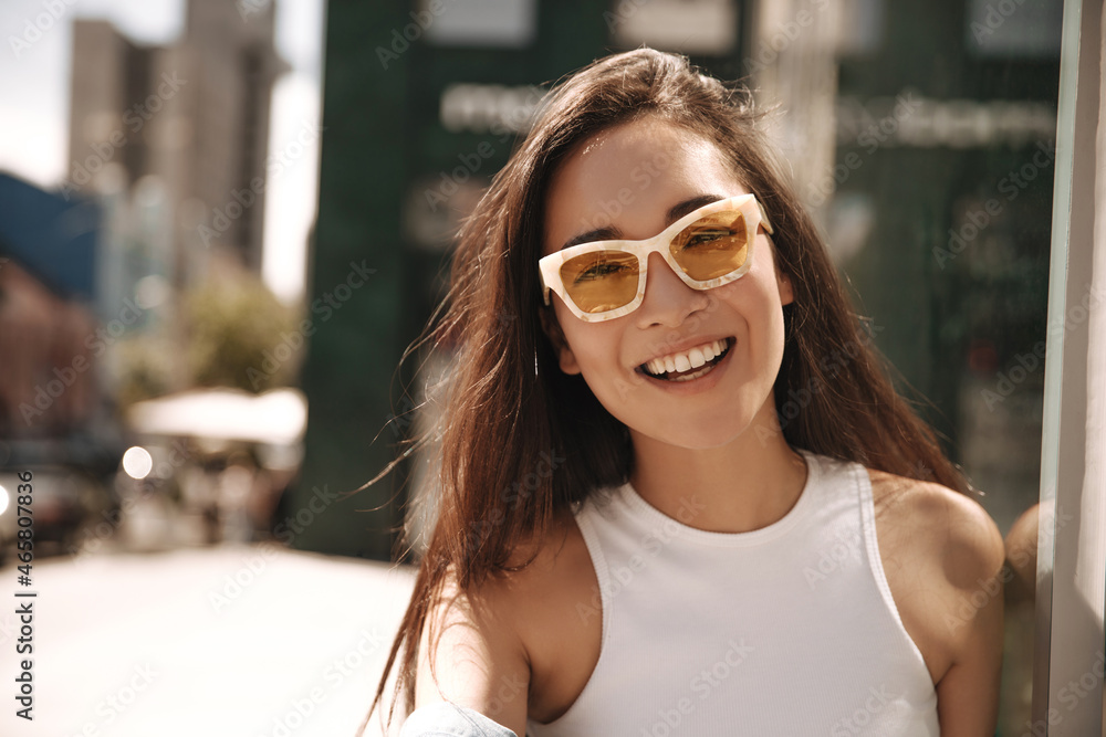 Close-up image of nice-looking young asian girl in sunglasses outdoors. Cheerful brunette smiles broadly with teeth and looks into camera. Summer vacation concept.