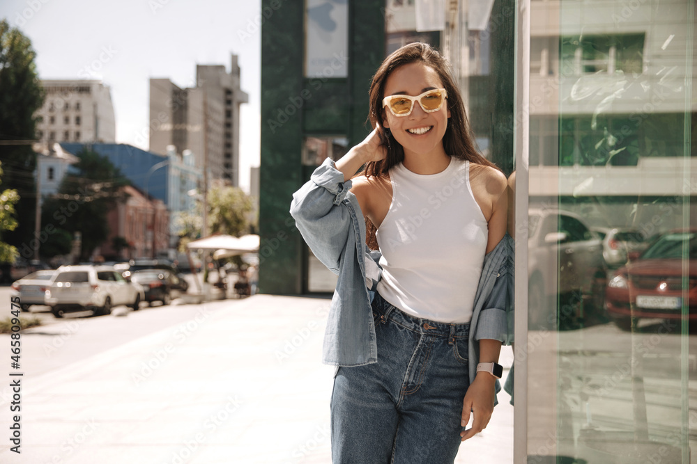 Summer sunny image of young stylish hipster woman standing near wall of panoramic building. Brunette in cute light denim clothes is resting and enjoying weekend.