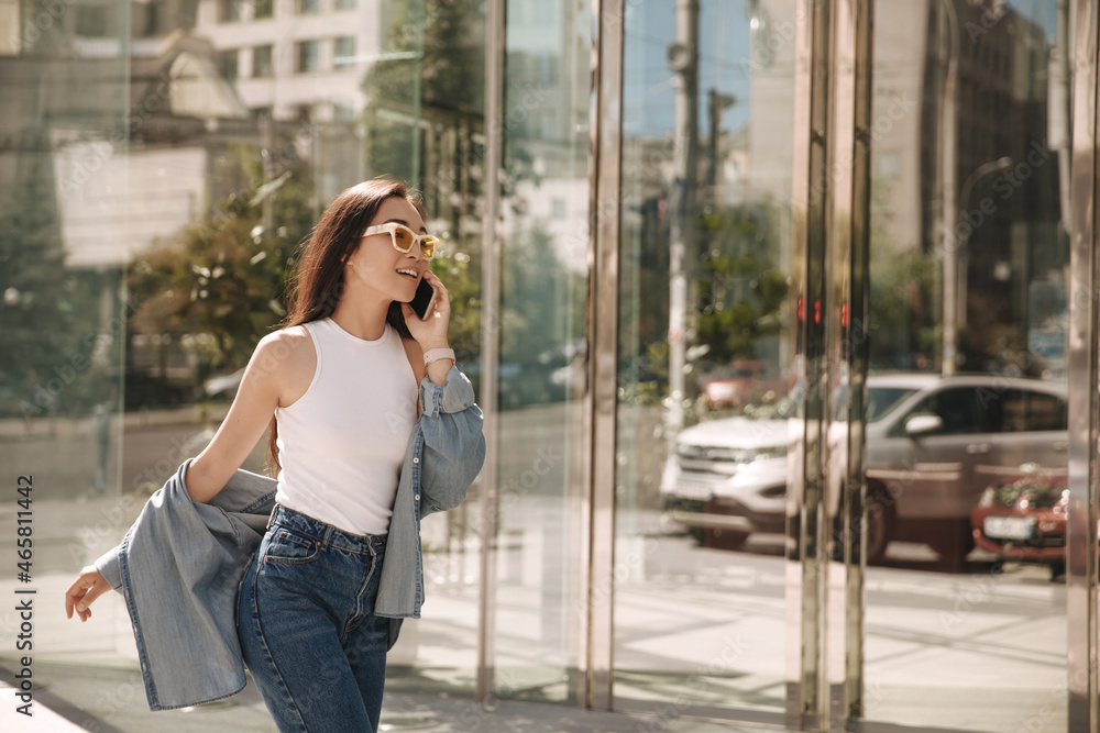 Fototapeta premium Asian woman walks in flying gait along panoramic windows of office. Brunette in sunglasses talking on phone with relatives. Dressed in white blouse, jeans and shirt.