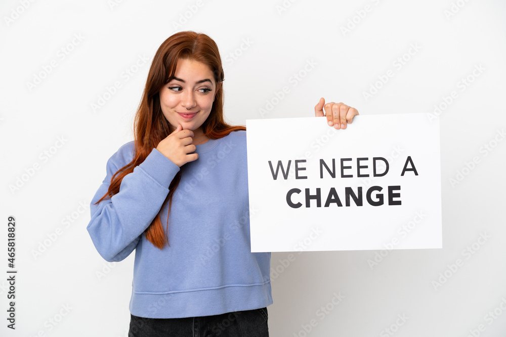Young redhead woman isolated on white background holding a placard with text We Need a Change and thinking Young redhead woman isolated on white background holding a placard with text We Need a Change and thinking