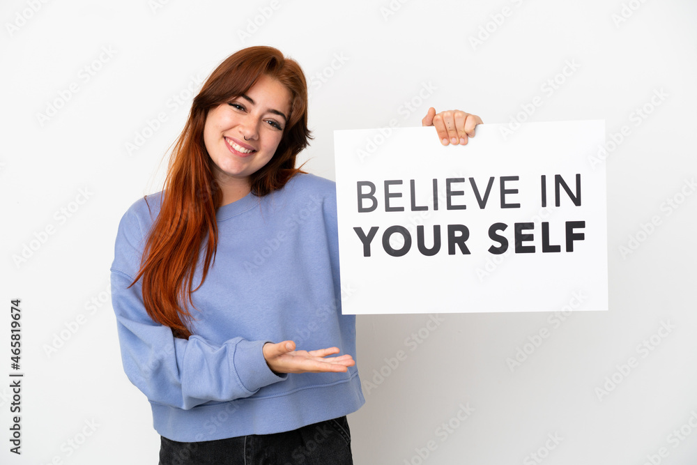 Young redhead woman isolated on white background holding a placard with text Believe In Your Self and pointing it Young redhead woman isolated on white background holding a placard with text Believe In Your Self and pointing it
