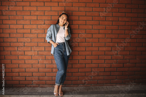 Joyful hipster young asian girl talking to friend through modern electronic gadget, standing near brick wall and looking to side. Dressed in light, light-colored clothes with glasses on her head.