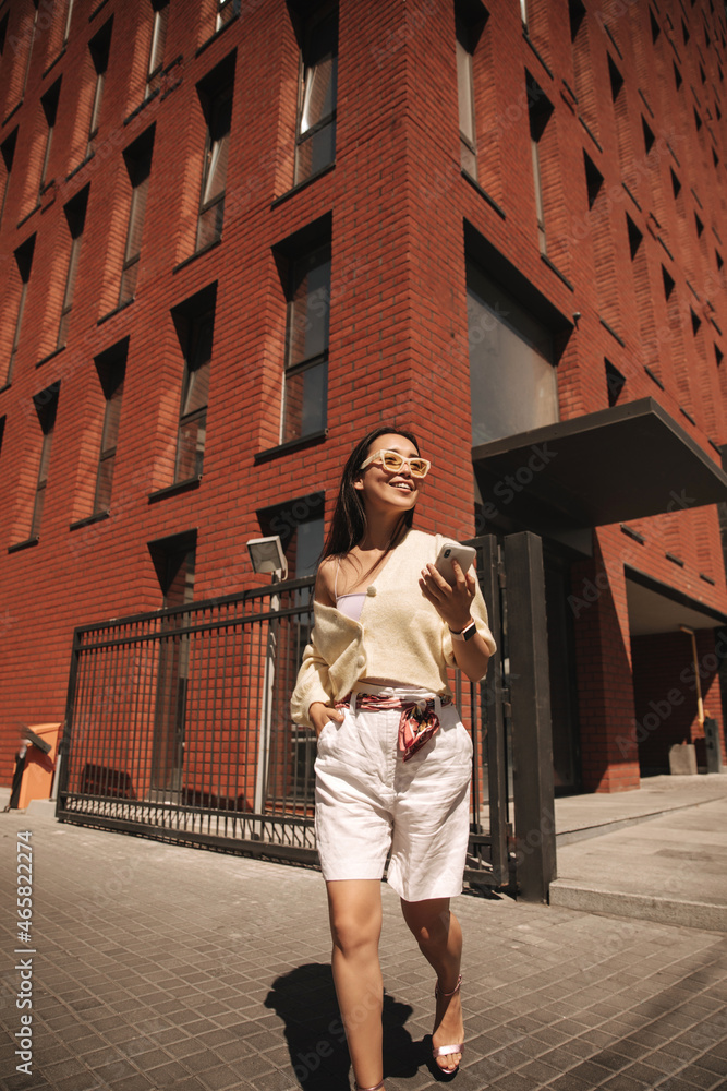 Stylish beautiful asian woman in shorts, sweater walks down street with smartphone in hand. Brunette in sunglasses. Elegant style, spring fashion trends. 