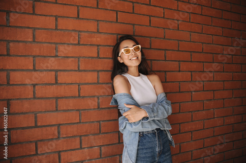 Pretty relaxed young asian woman hugs herself with her arms leaning against brick wall. Brunette with closed eyes wearing glasses smiles broadly in casual clothes.