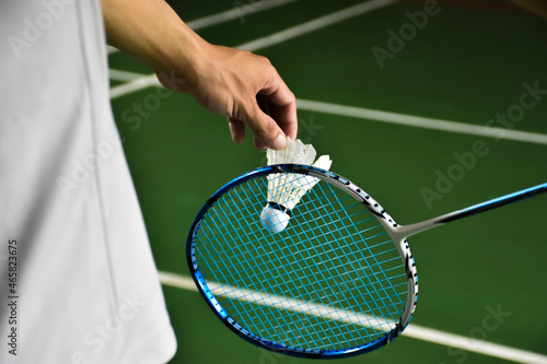 Badminton player is holding white badminton shuttlecock and badminton racket in front of the net before serving it over the net to another side of badminton court. Selective focus on white shuttlecock © Sophon_Nawit