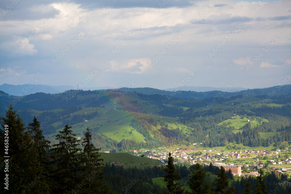 View on the village of Verkhovyna, spruce trees in the foreground, rainbow over mountains. Ukraine, Carpathians.