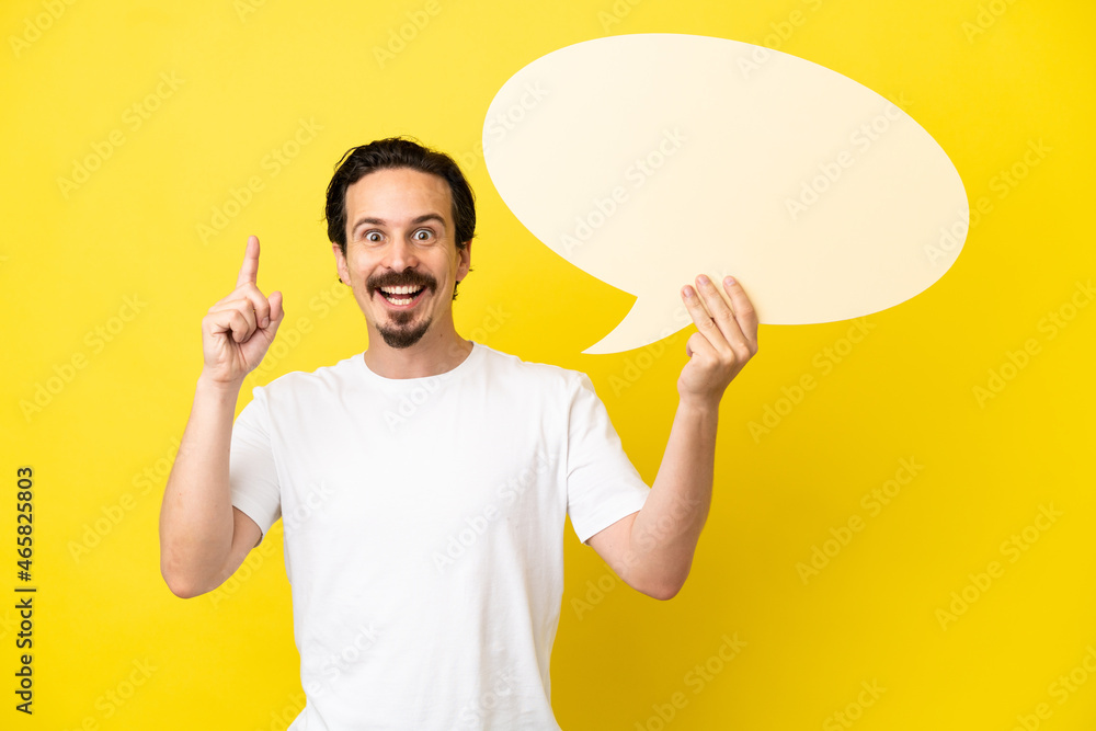Young caucasian man isolated on yellow background holding an empty speech bubble with surprised expression Young caucasian man isolated on yellow background holding an empty speech bubble with surprised expression