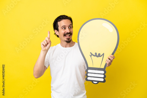 Young caucasian man isolated on yellow background holding a bulb icon and thinking