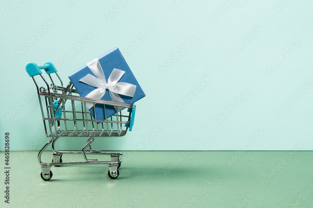 Beautiful blue gift box wrapped in ribbons in a shopping cart. Concept of online shopping around the world. Front view on a light background.