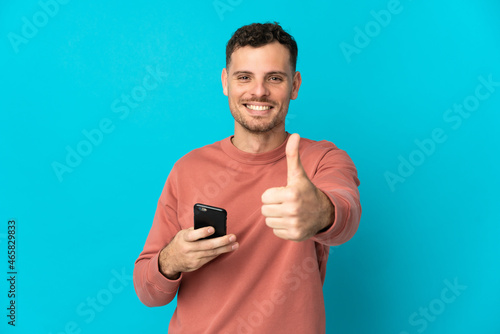 Young caucasian handsome man isolated on blue background using mobile phone while doing thumbs up