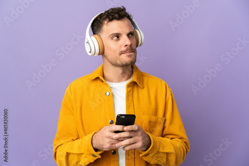 Young caucasian handsome man isolated on purple background listening music with a mobile and thinking © luismolinero