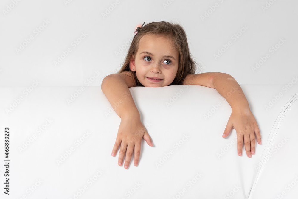 little blonde-haired, sympathetic emotional girl in a colored dress with blond hair leaned on the side of a former leather sofa on a white background, hung up his hands, looked. She is thoughtful little blonde-haired, sympathetic emotional girl in a colored dress with blond hair leaned on the side of a former leather sofa on a white background, hung up his hands, looked. She is thoughtful