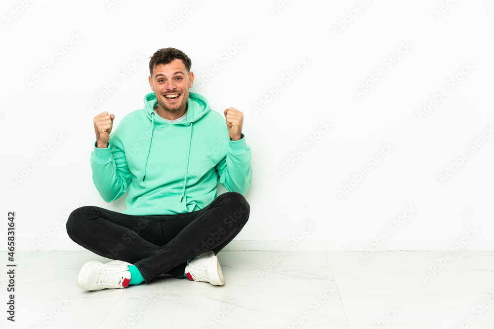Young handsome caucasian man sitting on the floor celebrating a victory in winner position Young handsome caucasian man sitting on the floor celebrating a victory in winner position