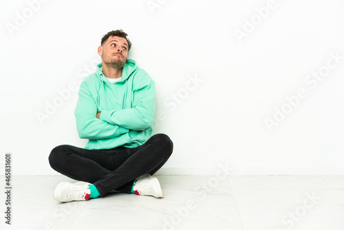 Young handsome caucasian man sitting on the floor making doubts gesture while lifting the shoulders