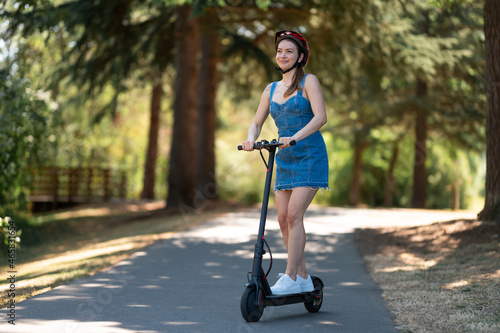 A nice girl in a protective helmet and denim dress rides an electric scooter in the park. Clear sunny day, beautiful nature. Eco-friendly transport, outdoor activities, sports.