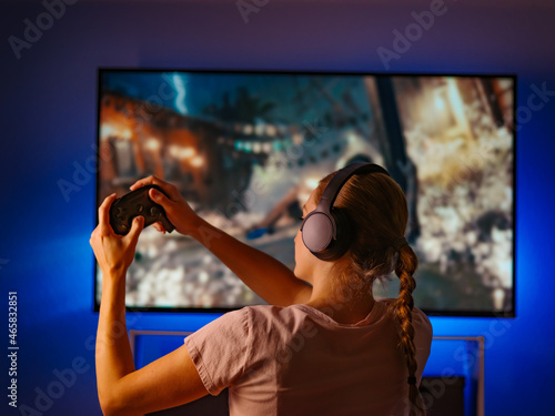 A girl with headphones and a joystick in her hands sits in front of a large monitor, plays a video game. Evening, insomnia, day off, vacation, gamer, gambling business. Shooting from the back.