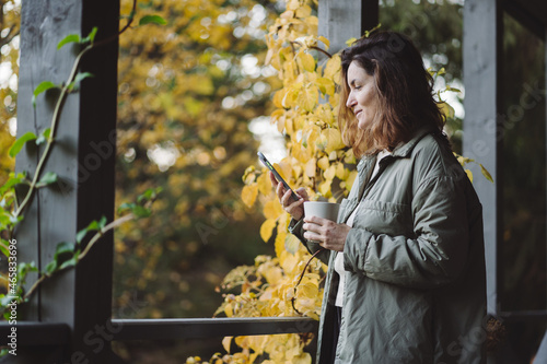  A young woman is drinking coffee and using the phone on the terrace in late autumn.
