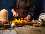 The chef is dicing an orange pumpkin for American Pumpkin Pie on a cutting board. Ingredients, decor. Wooden texture, blue background. Cooking process. cookbook, culinary blog, instagram.
